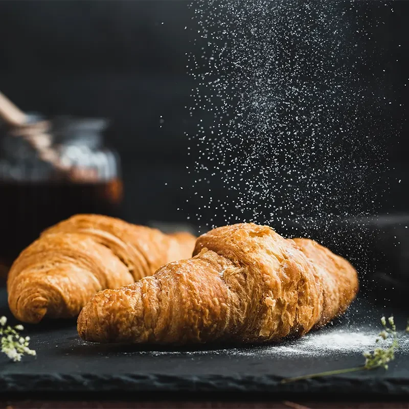Freshly baked golden croissants being dusted with powdered sugar, made using high-quality instant dry yeast for rich flavor and perfect rise in pastry baking.